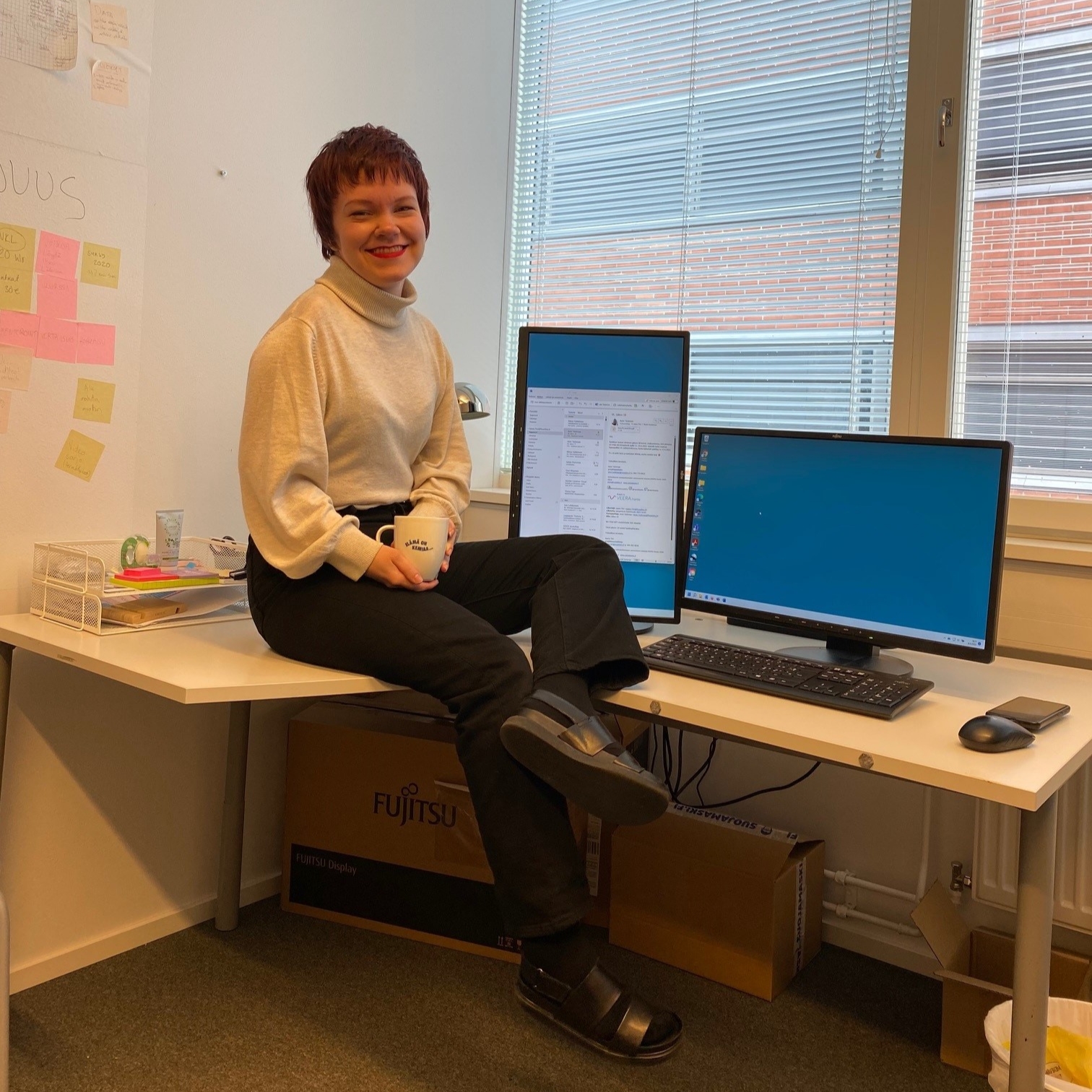 jaana_laksiaiset Jaanan valokuva viimeisestä työpäivästä. Jaana istuu työpöydällä kahvikuppi kädessä.