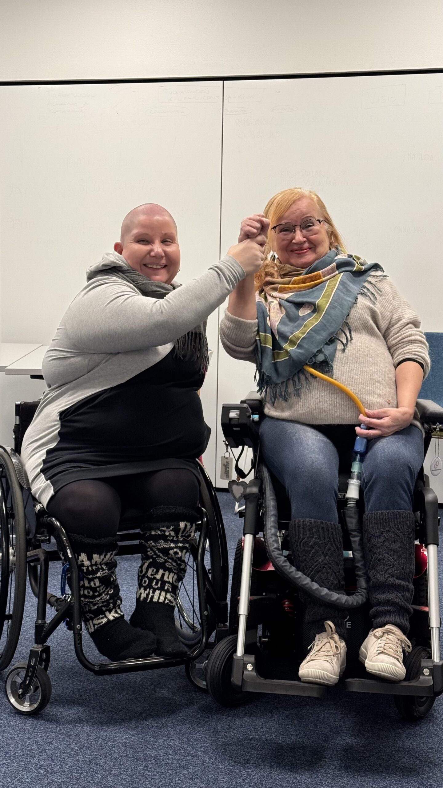 Valokuva Annesta ja Ritasta. Molemmat naiset istuvat pyörätuolissa, Anne manuaalipyörätuolissa ja Rita sähköpyörätuolissa. Anne ja Rita nostavat yhdessä kättä ylös.